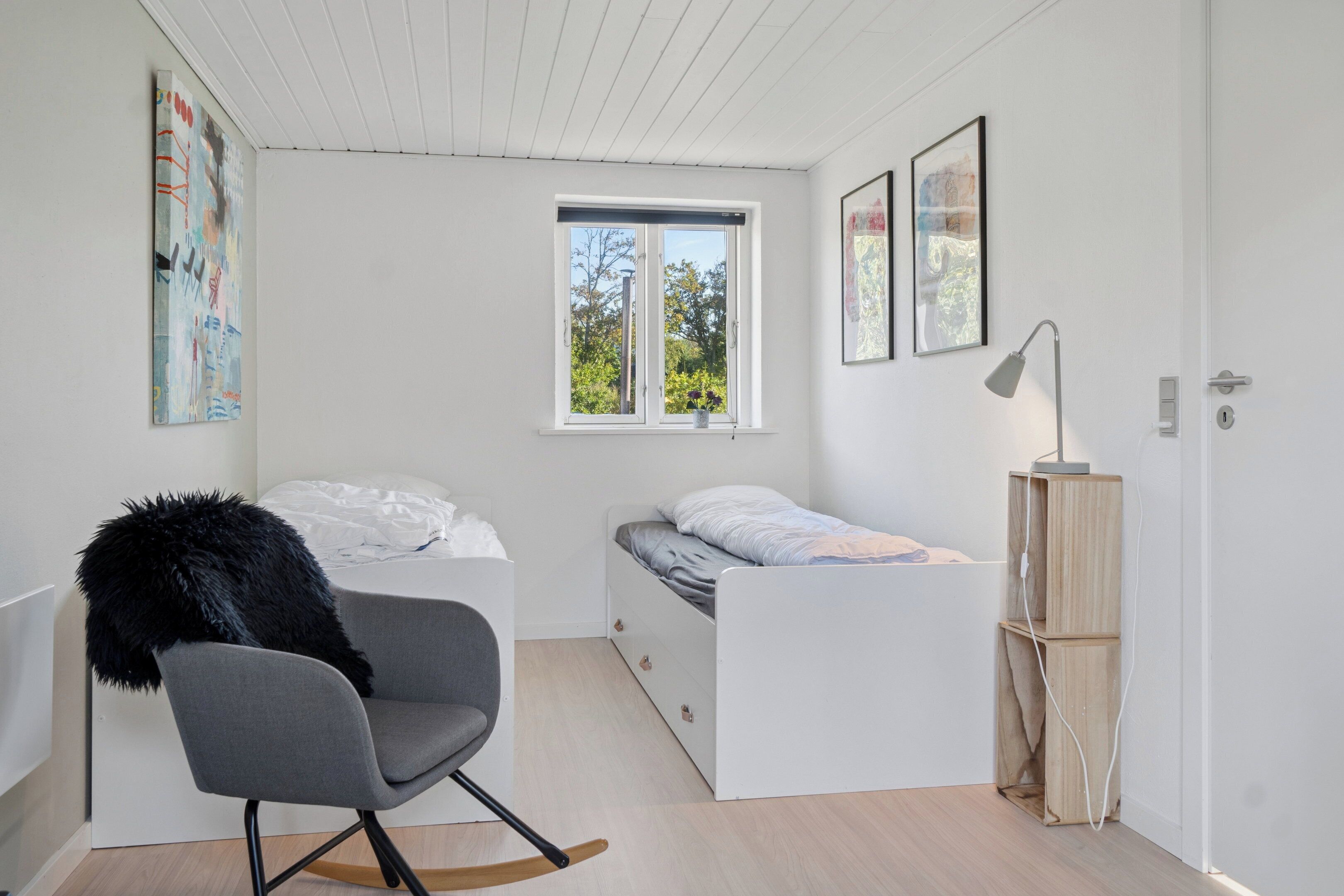 A compact bedroom with a bunk bed, a rocking chair, a bedside table with a lamp, and a window with a view of greenery.