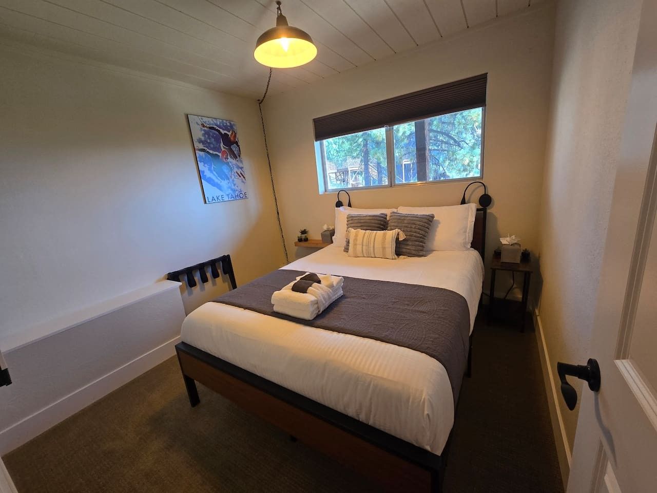 A neatly made bed with pillows and a blanket, a bedside table with a lamp, and a window with a view of trees.
