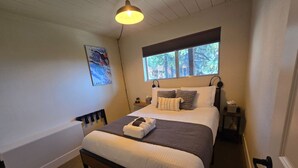 Een net opgemaakt bed met kussens en dekbed, een nachtkastje met een lamp en een raam met uitzicht op bomen.