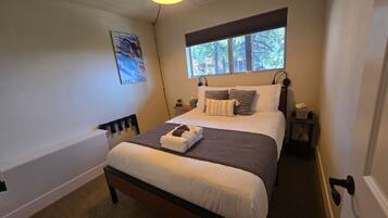 A neatly made bed with pillows and a blanket, a bedside table with a lamp, and a window with a view of trees.