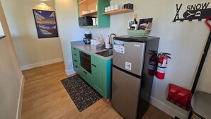 Een compacte keuken met een koelkast, magnetron en een klein spoelgedeelte. Boven het aanrecht bevinden zich houten planken en aan de muur is een brandblusser bevestigd.
