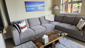 Een woonkamer met een grijze bank, een houten salontafel en een schilderij van een landschap aan de muur.