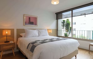 A hotel room with a large bed, two bedside lamps, a framed picture on the wall, and a view of the outside through a large window. - w* | Amazing 2BR with Balcony in Prime Miraflores (Miraflores)