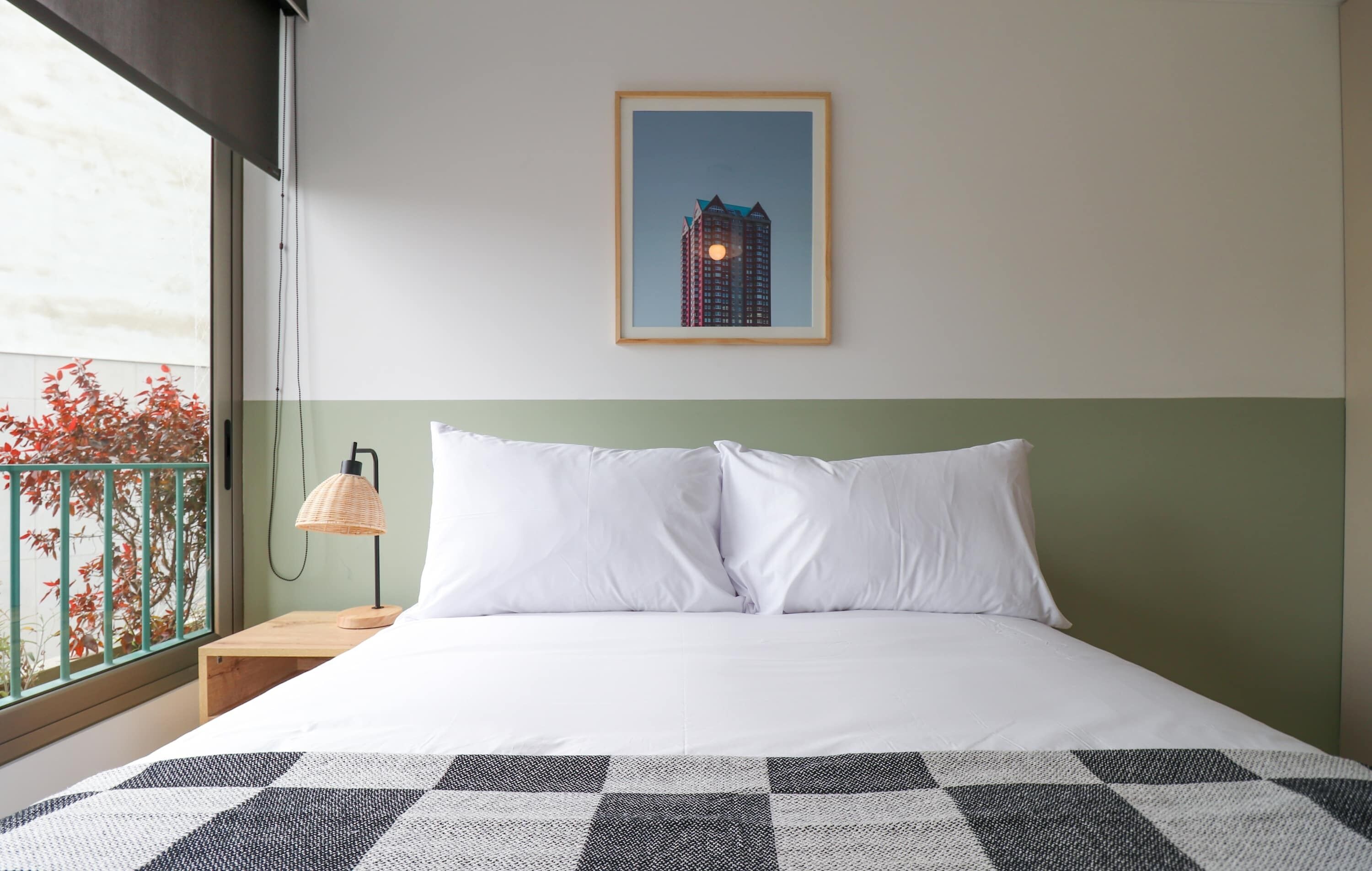 A hotel room with a bed, a nightstand, a lamp, a framed picture on the wall, and a view of the outside.