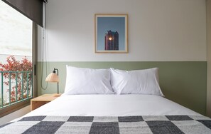 A hotel room with a bed, a nightstand, a lamp, a framed picture on the wall, and a view of the outside. - w* | Amazing 2BR with Balcony in Prime Miraflores (Miraflores)