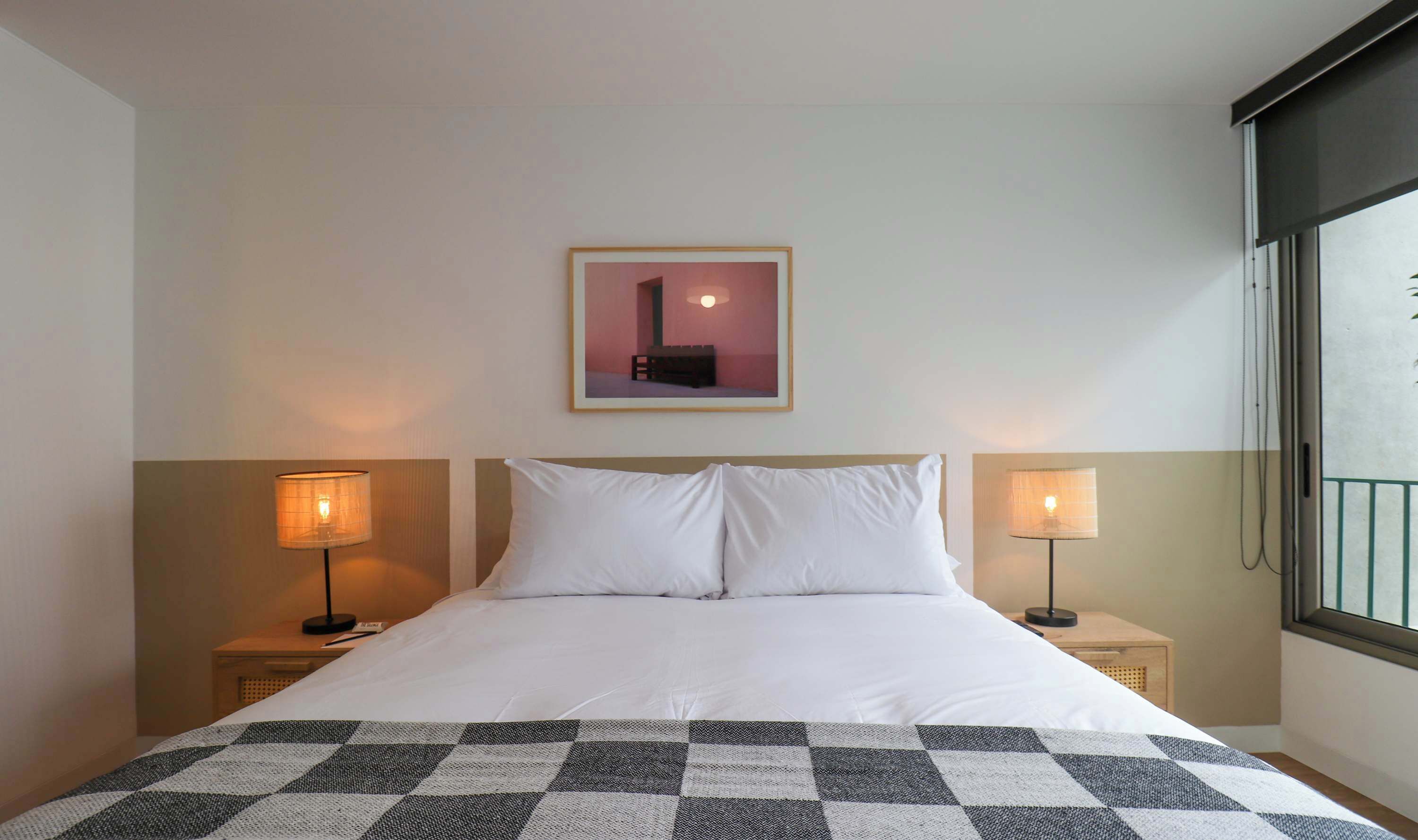 A hotel room with a bed, two bedside lamps, a framed picture on the wall, and a window with a view.