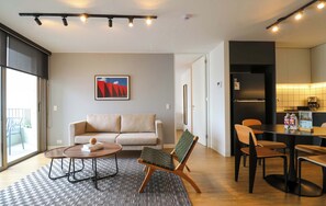 A modern living room with a sofa, a coffee table, and a chair. There is a dining area with a table and chairs. A kitchen is visible in the background. - w* | Upscale 2BR with Balcony in Prime Miraflores (Miraflores)