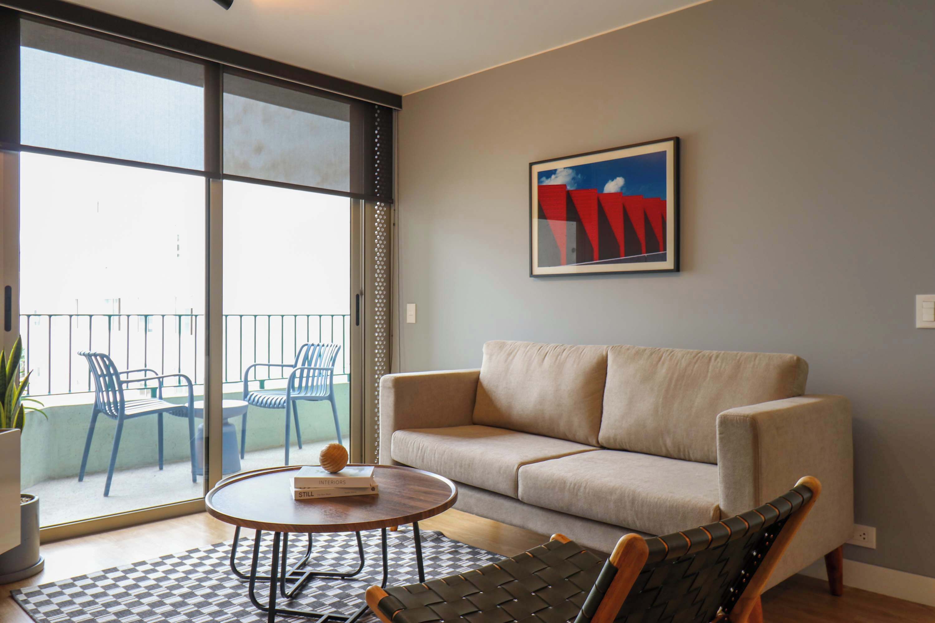 A living room with a sofa, a round coffee table, and a chair. There is a large window with a view of outdoor chairs and a balcony.
