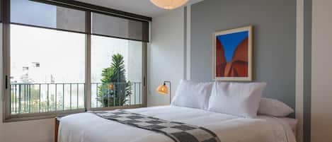 A bedroom with a large window, a bed with white bedding, a hanging light fixture, and a painting on the wall.