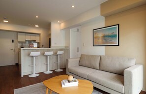 A modern living room with a sofa, a coffee table with a book, and a kitchen area with bar stools. - w* | Enchanting 1BR w/ Balcony in Prime Miraflores (Miraflores)