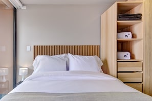 Et hotelværelse med en seng, et træsengegærde, et natbord med en lampe og en garderobe med foldede håndklæder.