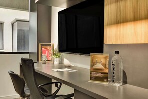Ett modernt hotellrum med ett skrivbord, en stol, en krukväxt, en inramad tavla, en flaska och en reklamaffisch.