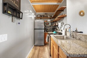 A compact kitchen with a stainless steel refrigerator, a stove, and a sink. There are wooden cabinets and a granite countertop.