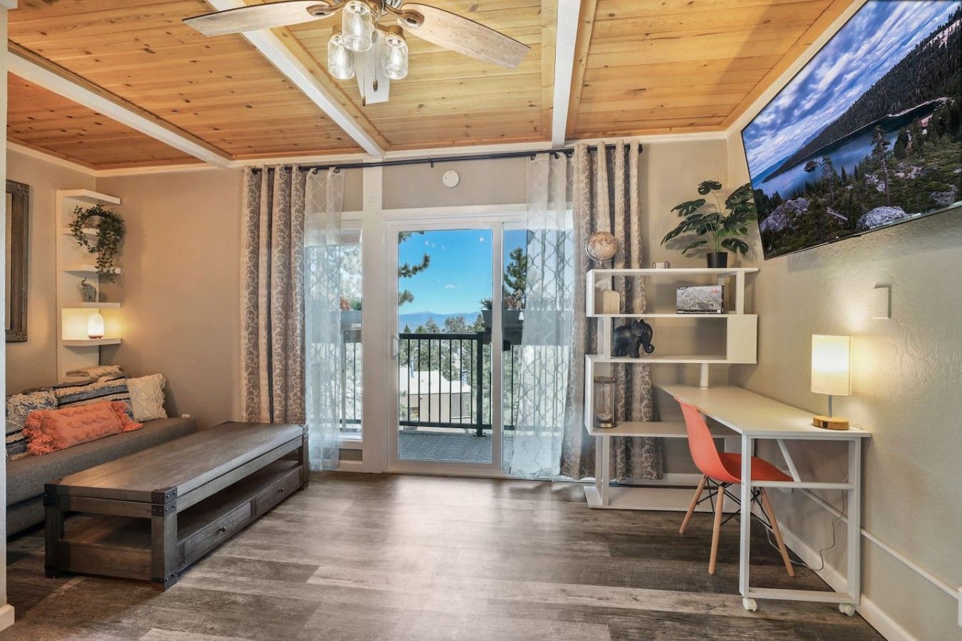 A room with a wooden ceiling, a sofa, a desk with a chair, a bookshelf, and a view through a glass door.