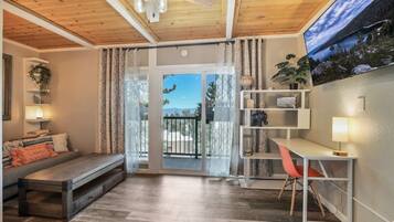 A room with a wooden ceiling, a sofa, a desk with a chair, a bookshelf, and a view through a glass door.