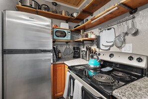 A compact kitchen with a stainless steel refrigerator, a stove with four burners, and a microwave. There are wooden shelves above the stove holding various kitchen items. - Couple’s King Suite | Sauna & Balcony | Sleeps 3 (Stateline)