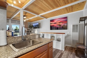 A kitchen with granite countertops, stainless steel sink, wooden cabinets, a wooden ceiling, and a TV mounted on the wall.