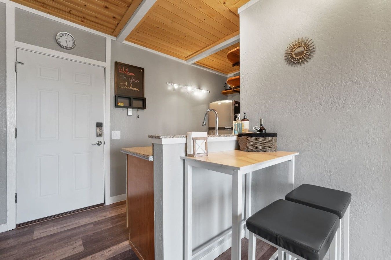 A kitchen with a wooden ceiling, a white door, a welcome sign, a sink, and a countertop.