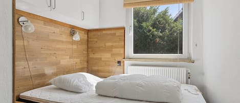 A bedroom with a bed, wooden headboard, two bedside lamps, a window with a view of trees, and a radiator.