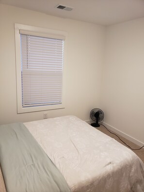 A small, unadorned room with a single bed, a window with blinds, a wall-mounted fan, and a power outlet. - Charming bedroom in fantastic Spartanburg with cool AC (Spartanburg)