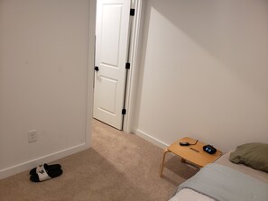 A bedroom with a bed, a nightstand, a wooden table, a pair of slippers, and a door leading to another room. - Charming bedroom in fantastic Spartanburg with cool AC (Spartanburg)