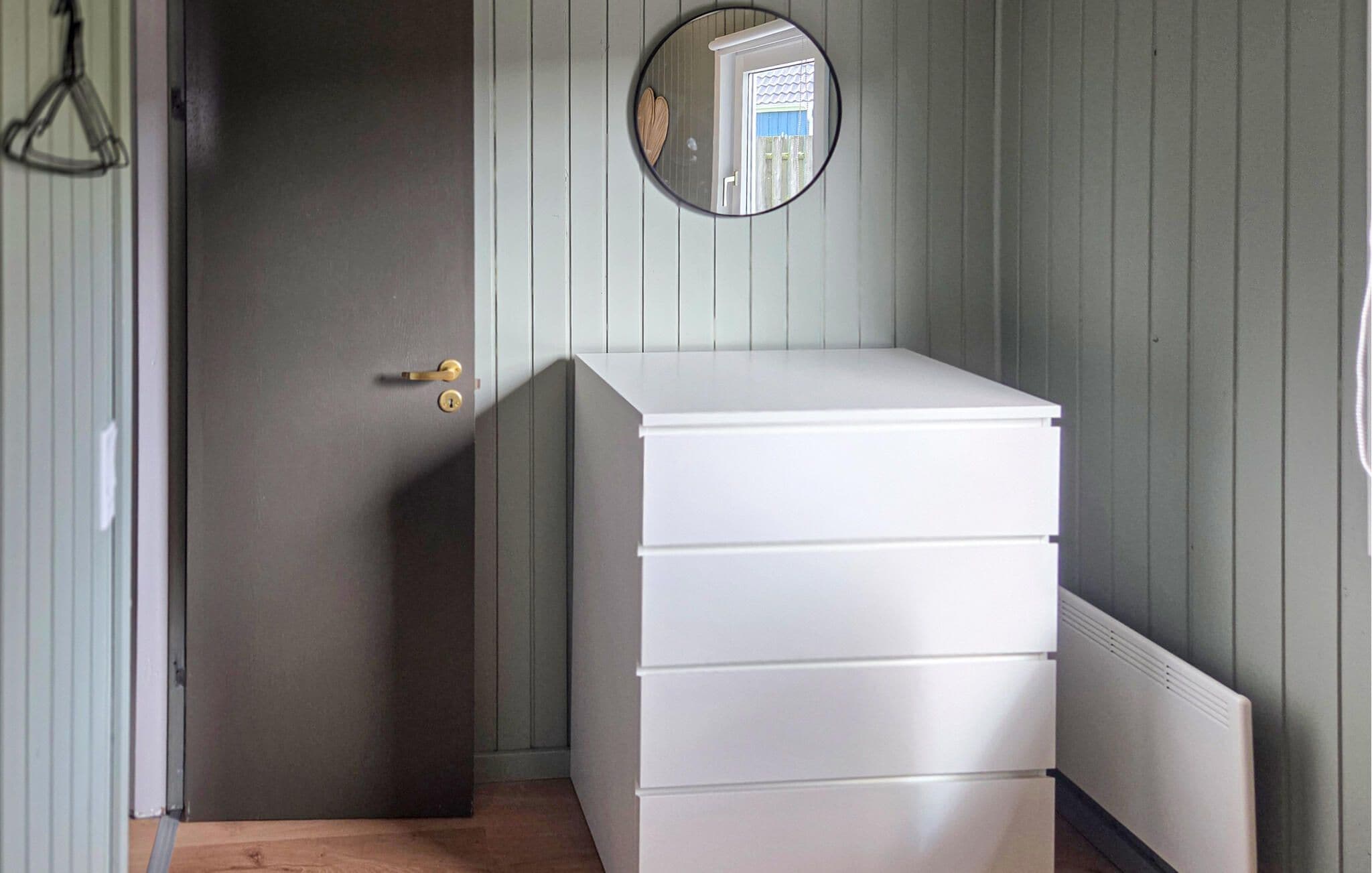Una habitación con un aparador blanco, una puerta con picaporte de latón y un espejo redondo en la pared.