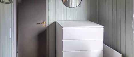 Une pièce avec une commode blanche, une porte dotée d’une poignée en laiton et un miroir rond accroché au mur.