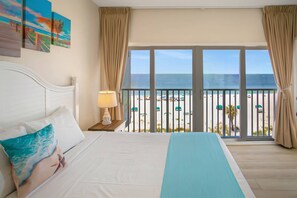 A hotel room with a bed, a nightstand, a lamp, and a view of the beach. - Three Bedroom Beachfront | Top Floor | Panoramic Gulf Views | Heated Pool, Tiki Bar & Beach Access (Treasure Island)