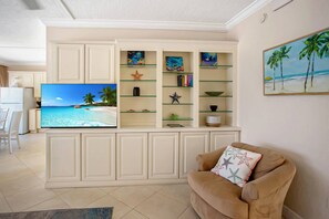 A living room with a flat-screen TV, a beige sofa with starfish pillows, a glass display unit with decorative items, and a painting of a beach scene on the wall. - Three Bedroom Beachfront | Top Floor | Panoramic Gulf Views | Heated Pool, Tiki Bar & Beach Access (Treasure Island)