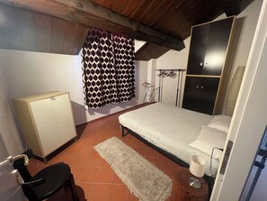 A bedroom with a bed, a wardrobe, a chair, a radiator, and a patterned curtain. - Badia del Borgo: A Home in the Millennium-Old Abbey (Toscana)