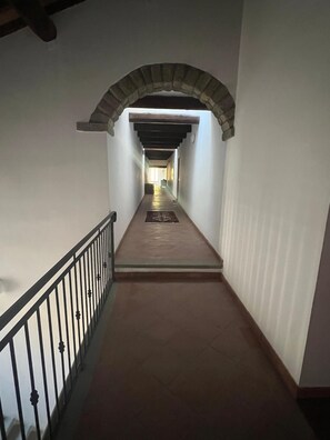 Un couloir étroit au sol carrelé, avec une rambarde métallique d’un côté et une arche en pierre au fond.