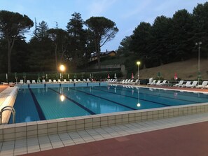 A swimming pool with multiple lanes, surrounded by lounge chairs and umbrellas, with trees and a building in the background. - Badia del Borgo: A Home in the Millennium-Old Abbey (Toscana)