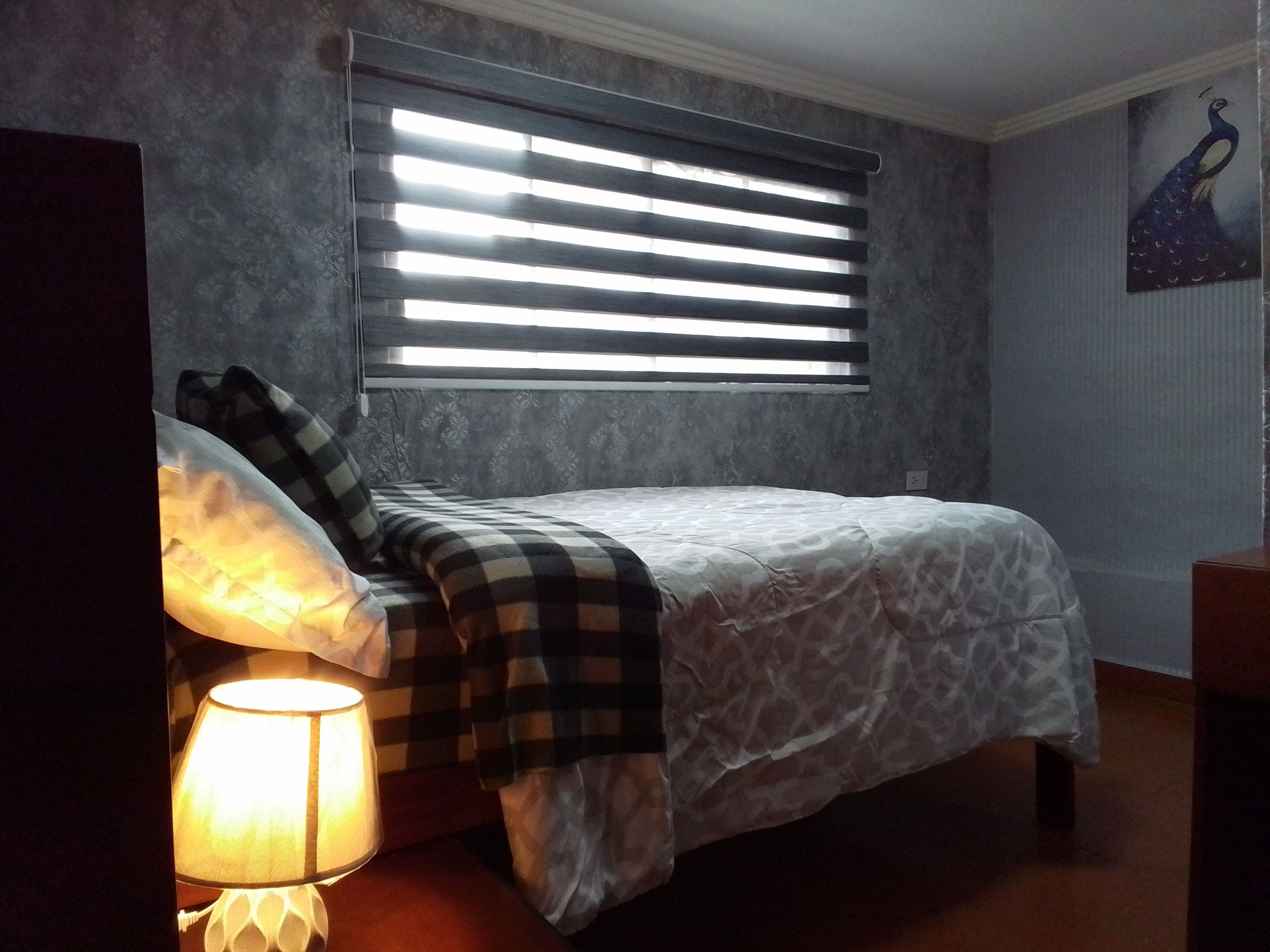 Un dormitorio con cama, una ventana con persianas, una lámpara y un cuadro en la pared.
