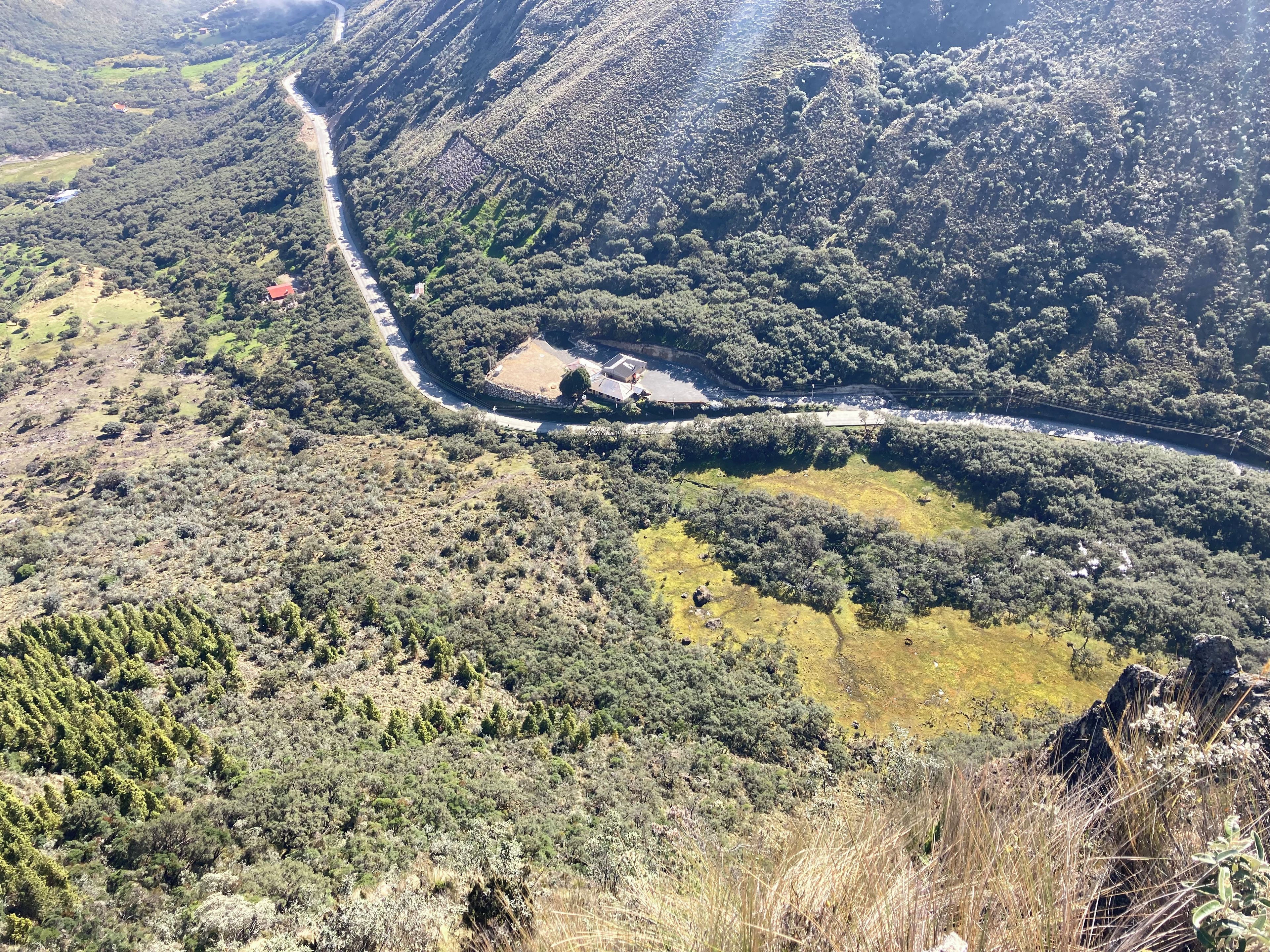 Un paisaje montañoso con un camino sinuoso, un pequeño edificio de techo rojo y vegetación densa.