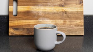 Una tazza bianca su un piano di lavoro scuro, con del vapore che sale, posizionata davanti a un tagliere di legno con un manico.