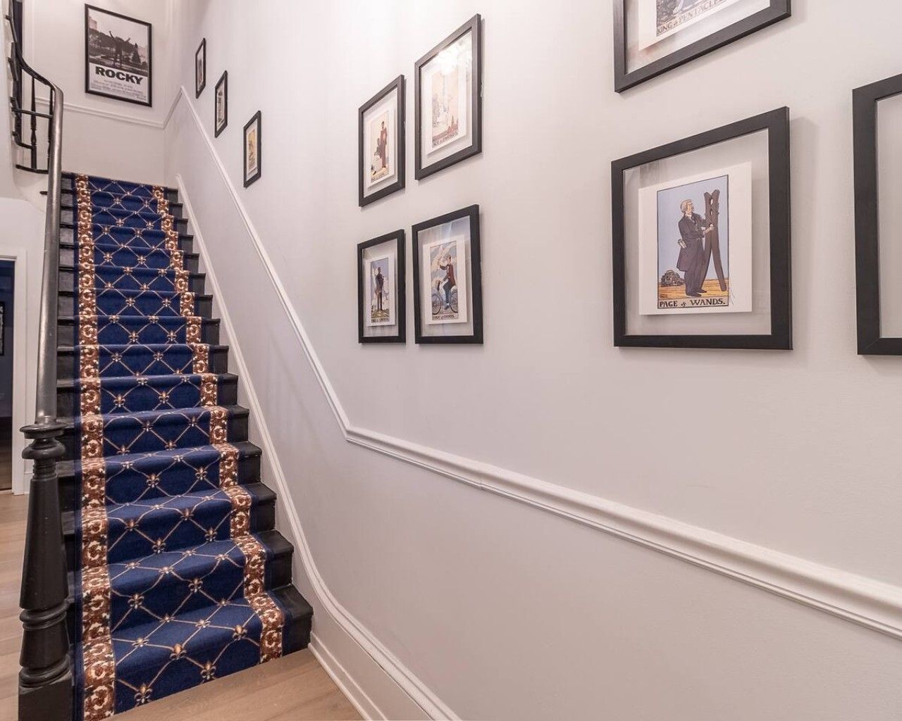 A staircase with a patterned carpet and a handrail, leading to a landing with framed pictures on the wall.