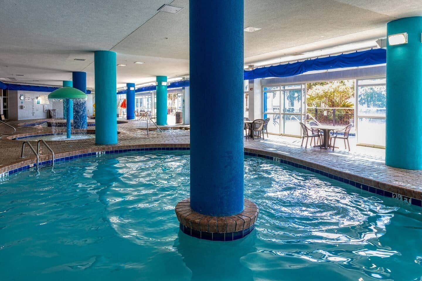 An indoor swimming pool with clear blue water, surrounded by blue pillars, and a seating area with tables and chairs.