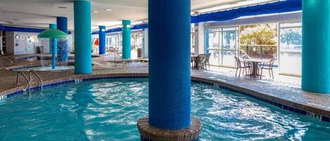 An indoor swimming pool with clear blue water, surrounded by blue pillars, and a seating area with tables and chairs.