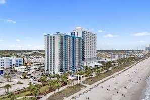 A coastal hotel with multiple floors, a beachside location, and a parking area with cars. - 1 Br Couples Beach Escape | Balcony + Resort Pools (Myrtle Beach)