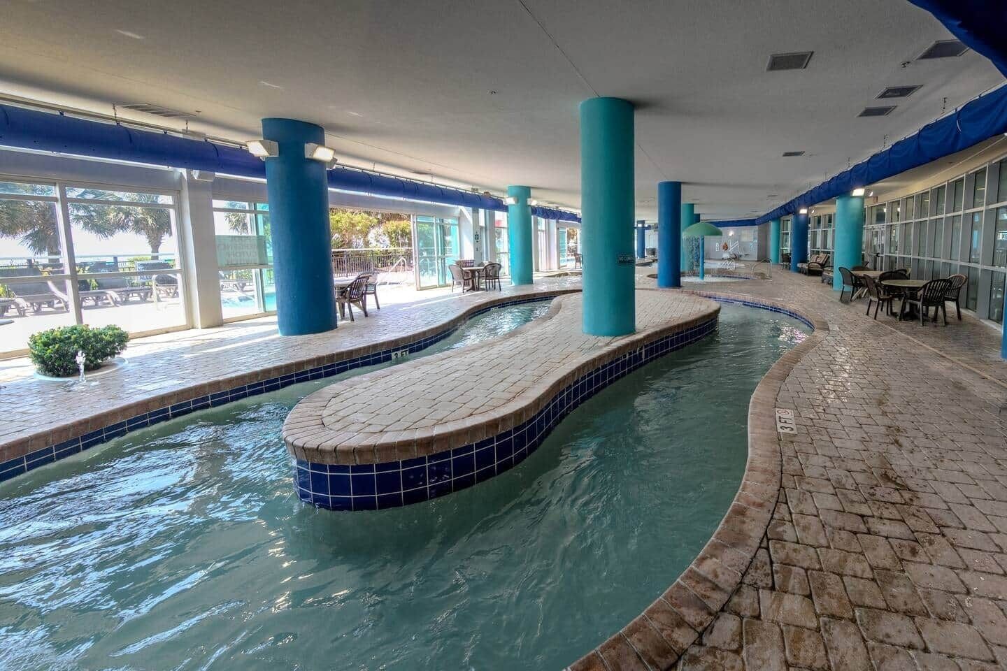 Una piscina interior con bordes de azulejos, pilares azules y zonas de estar al aire libre.