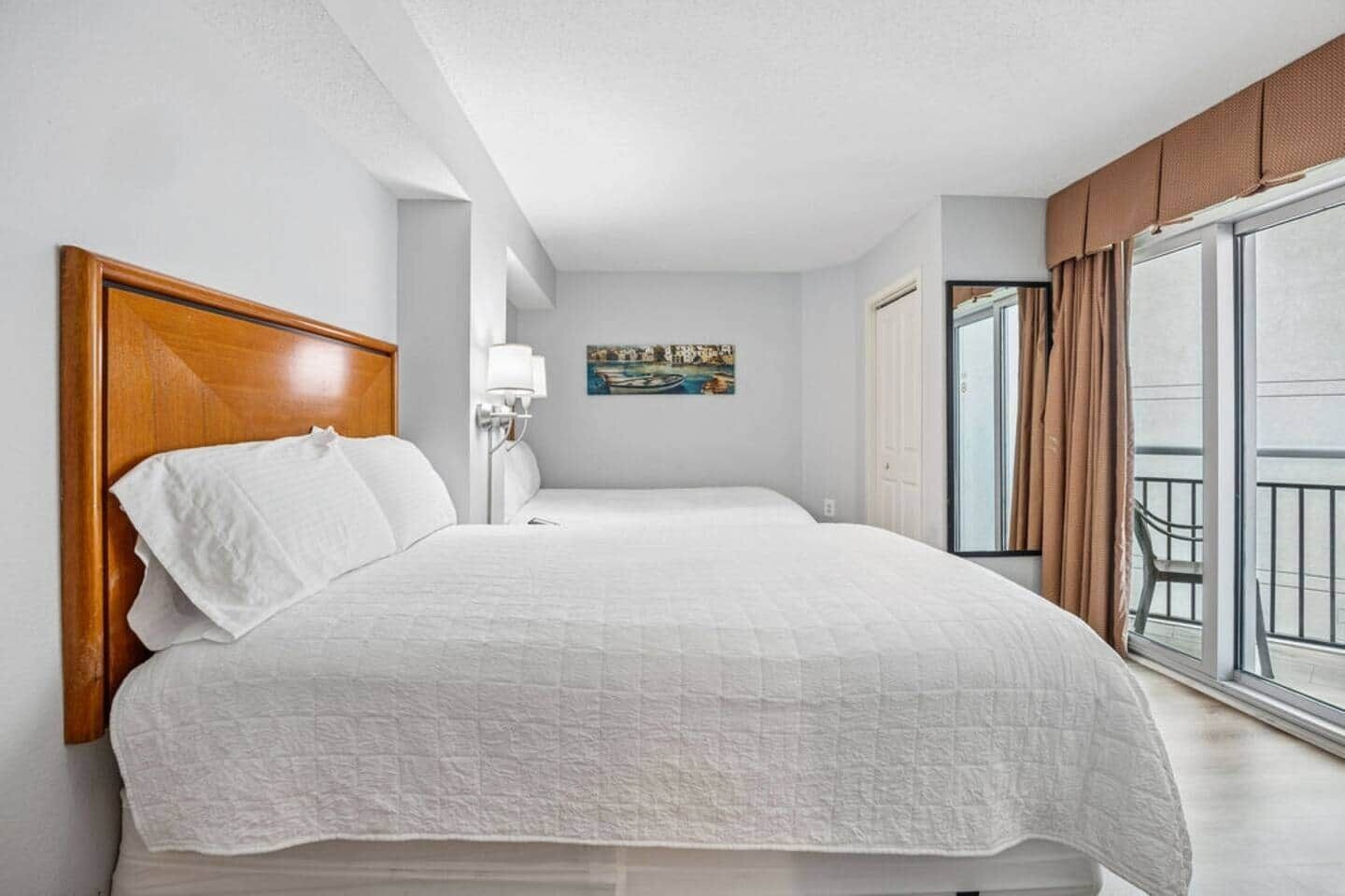 A hotel room with a large bed, a nightstand, a wall-mounted lamp, a painting, and a balcony with curtains.