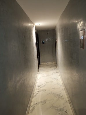 Corredor com piso de mármore e paredes metálicas, que leva a um elevador com placa indicando o número do andar.