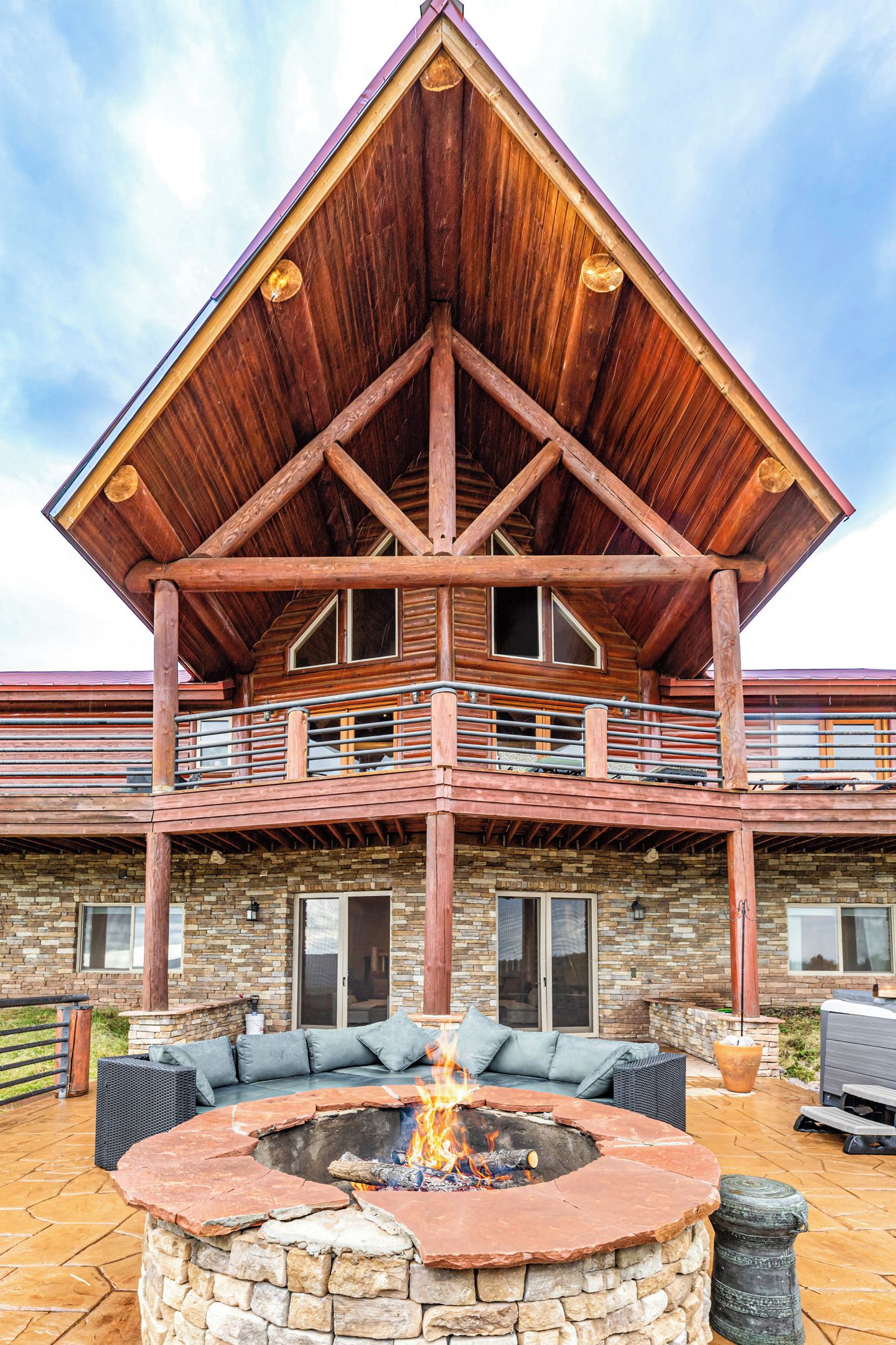A two-story log cabin with a stone foundation, a fire pit in the center, and a balcony with railings.