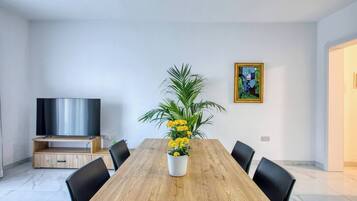Un comedor moderno con una mesa de madera, sillas negras, una planta en maceta con flores amarillas, un televisor de pantalla plana sobre un soporte y un cuadro en la pared.