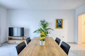 A modern dining area with a wooden table, black chairs, a potted plant with yellow flowers, a flat-screen TV on a stand, and a framed picture on the wall. - Modern Central 2-BR Apt. with Views of Valletta (Tal-Pietà)