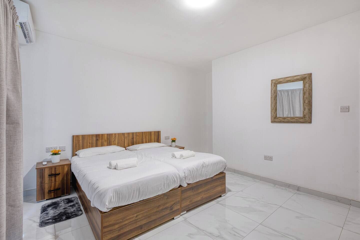 Habitación de hotel con una cama de madera, ropa de cama blanca, una mesita de noche con un florero y un espejo en la pared.