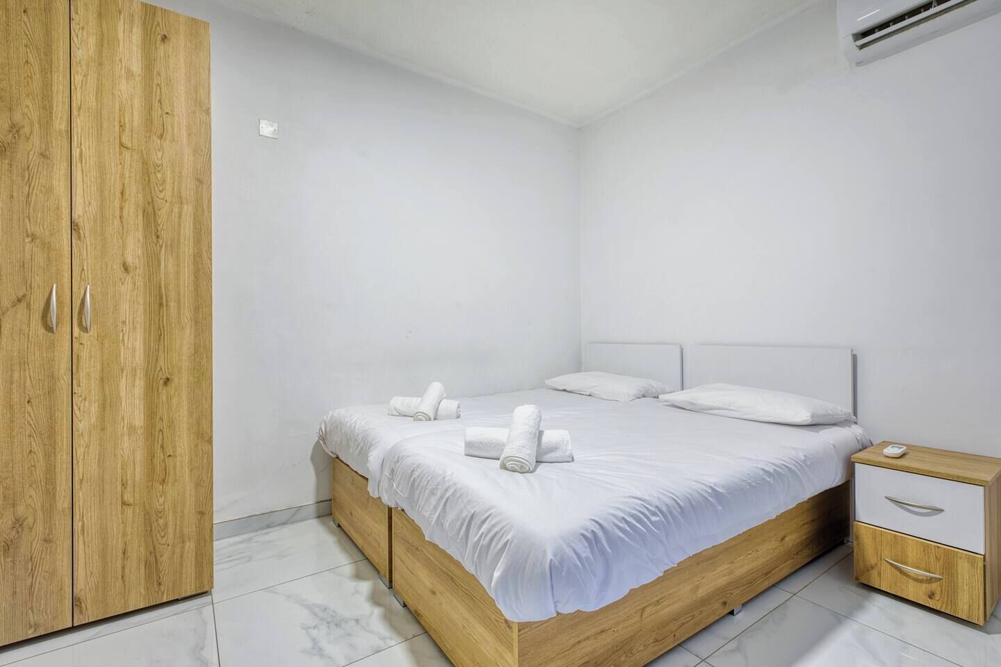 A bedroom with a wooden bed, white linens, a wooden wardrobe, and a bedside table with a lamp.