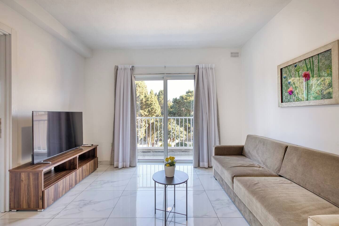 A modern living room with a flat-screen TV, wooden entertainment unit, a beige sofa, a small round table with a potted plant, and a balcony with curtains.
