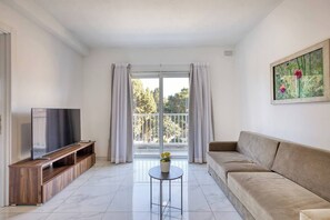 Un salon moderne avec un téléviseur à écran plat, un meuble de rangement en bois, un canapé beige, une petite table ronde agrémentée d’une plante en pot et un balcon avec des rideaux.