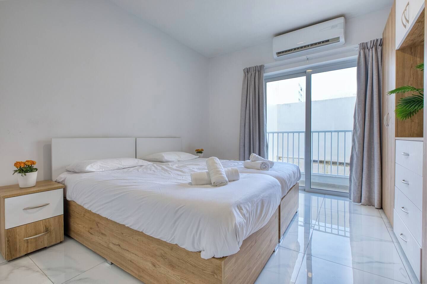 Quarto moderno com cama de madeira, mesa de cabeceira, cômoda e varanda com cortinas.